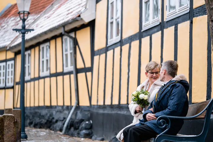 Brudepar på en bænk ved et bindingsværkshus